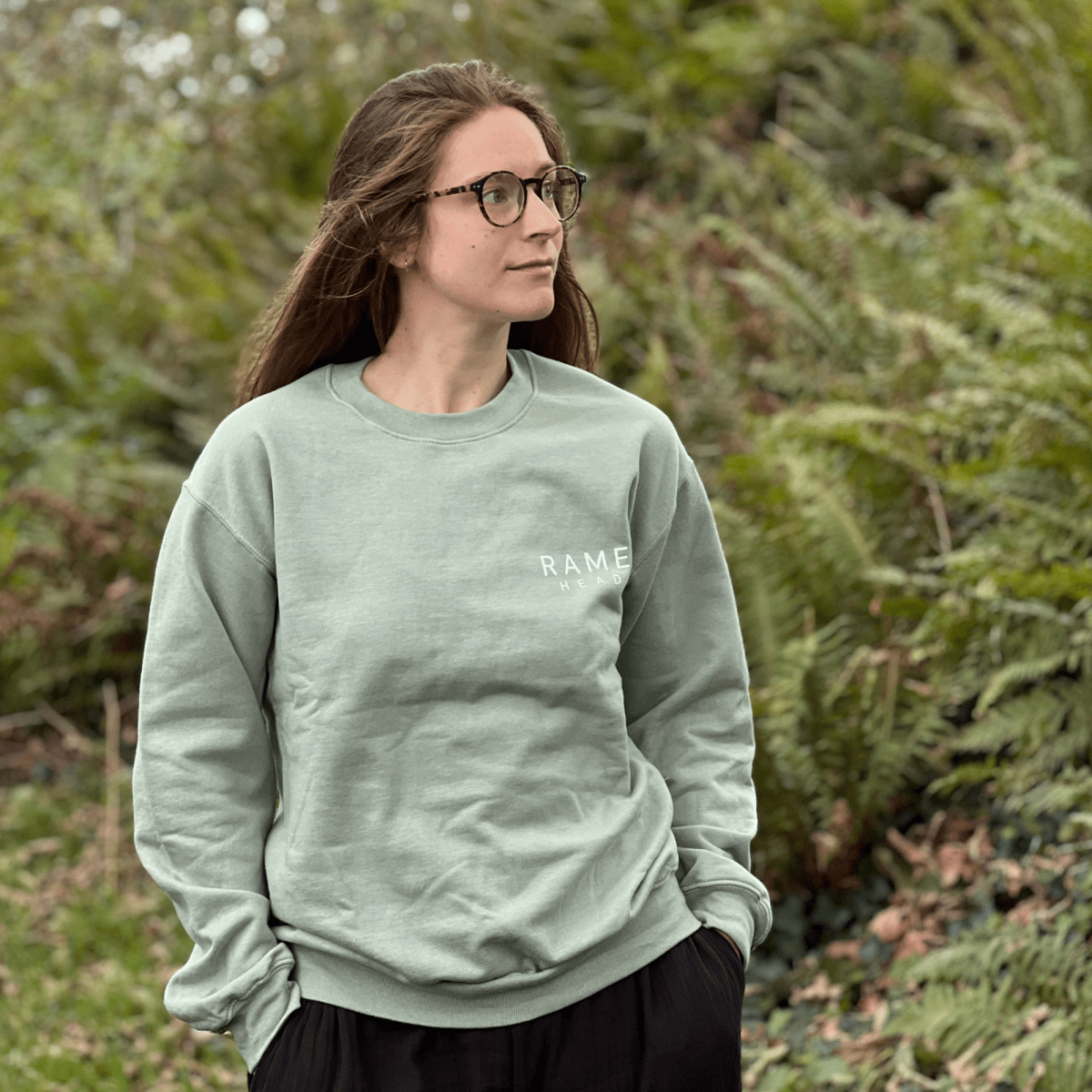 A woman with glasses and long brown hair wearing a sage green crewneck sweatshirt with 'RAME head' hand-screen printed in white on the chest, standing outdoors in front of a blurred background of green ferns. solf by Helen Round