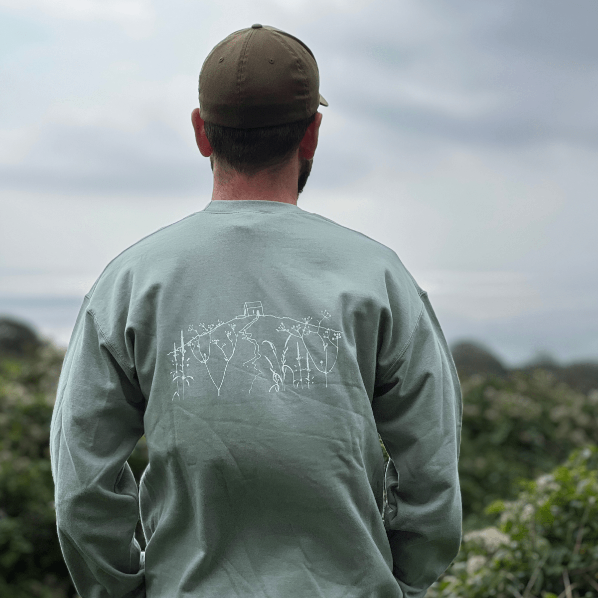 A man viewed from behind wearing a sage green long-sleeve shirt with a white line-art graphic on the back, paired with an olive green cap, set against a soft-focus outdoor background from Helen Round