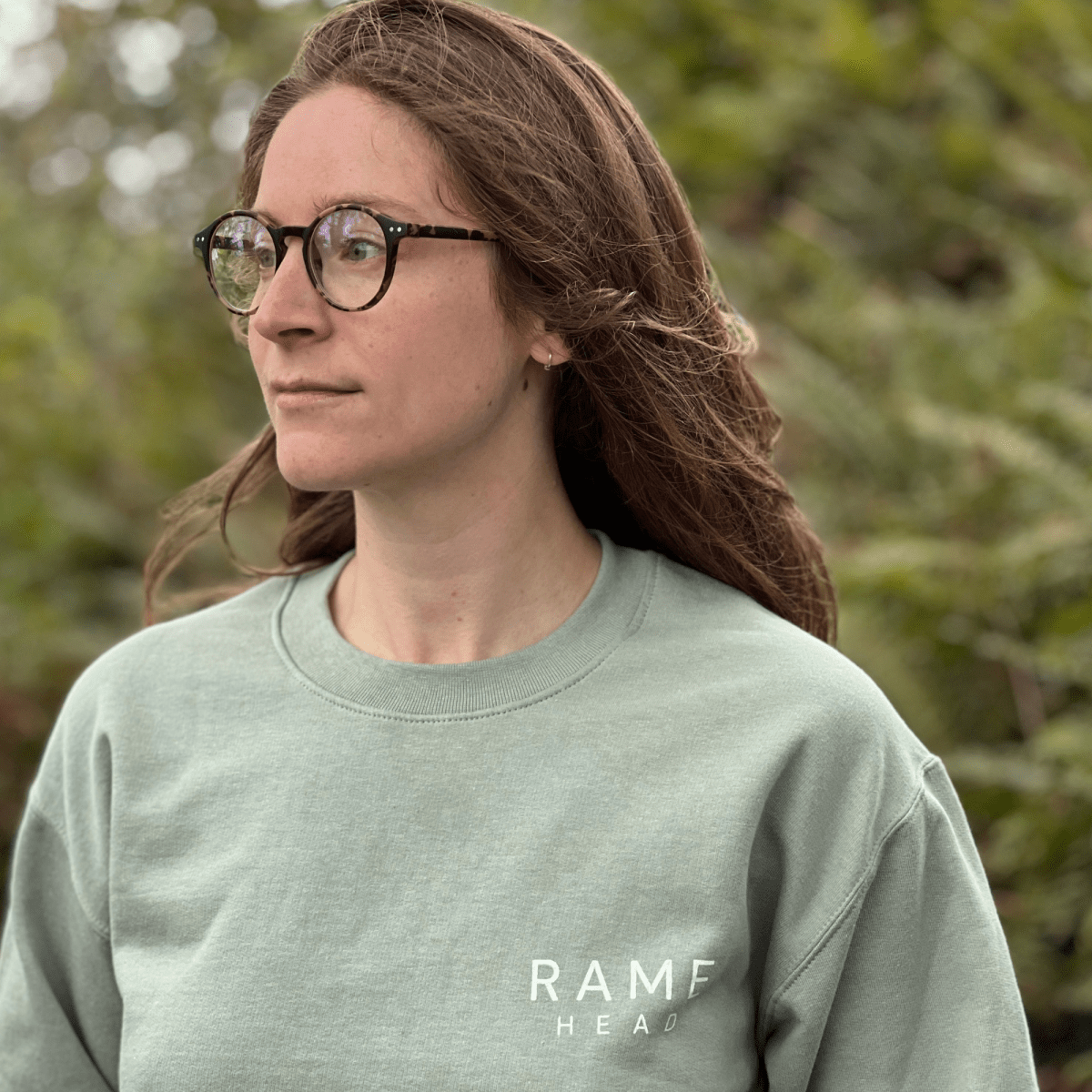 A woman with glasses and long brown hair wearing a sage green crewneck sweatshirt with 'RAME head' hand-screen printed in white on the chest, standing outdoors in front of a blurred background of green ferns. solf by Helen Round