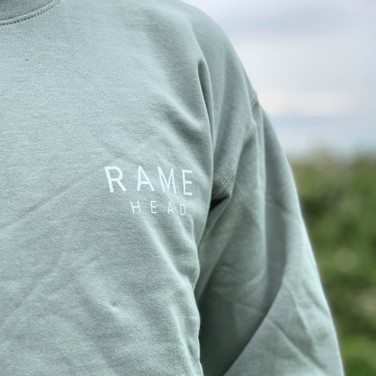 A close-up of a person wearing a sage green sweatshirt with 'RAME HEAD' printed in white on the chest, set against a blurred outdoor background.