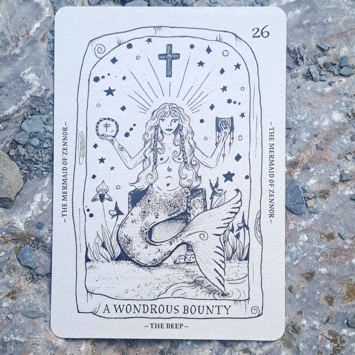 A tarot card titled 'A Wondrous Bounty' featuring an illustration of a mermaid, placed flat on a textured gray stone surface.