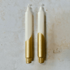 Two candles with white bodies and gold bases on a textured surface