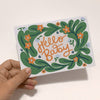 Hand holding a card with decorative text and green wreath design on a light background  sold by Helen Round