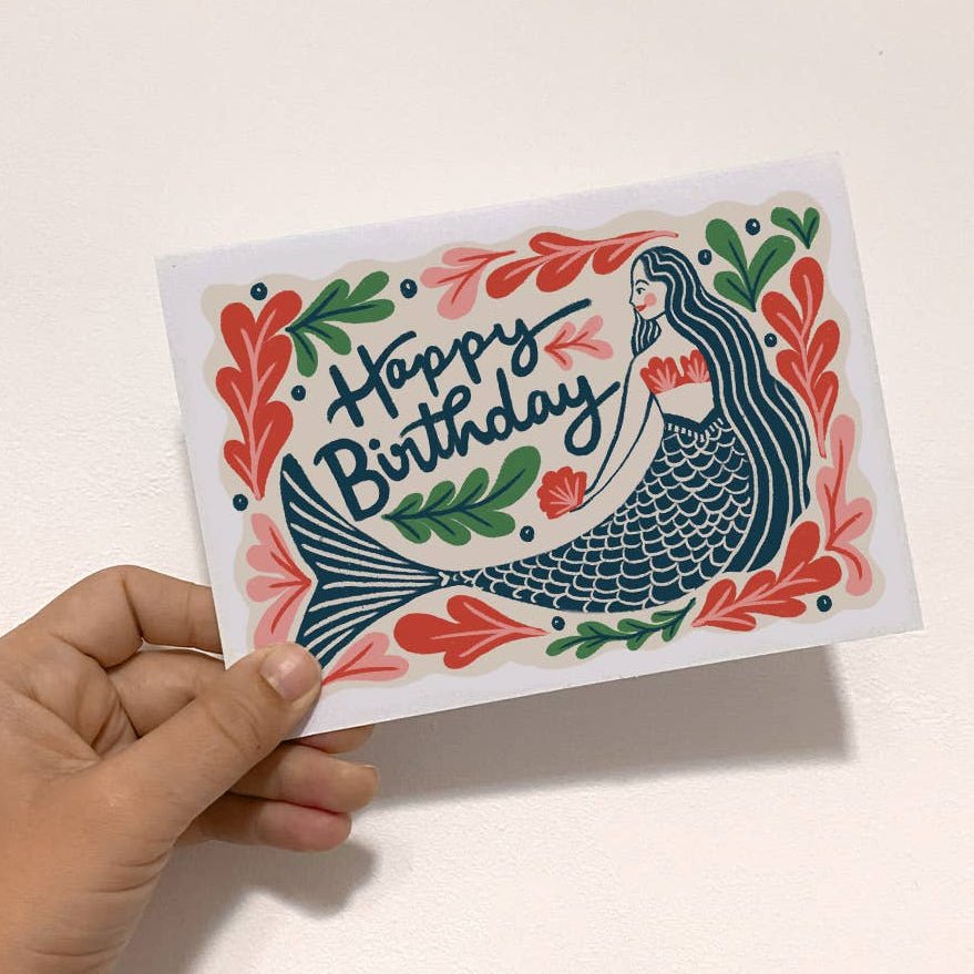 Hand holding a birthday card with mermaid design and 'Happy Birthday' text on a light background sold by Helen Round