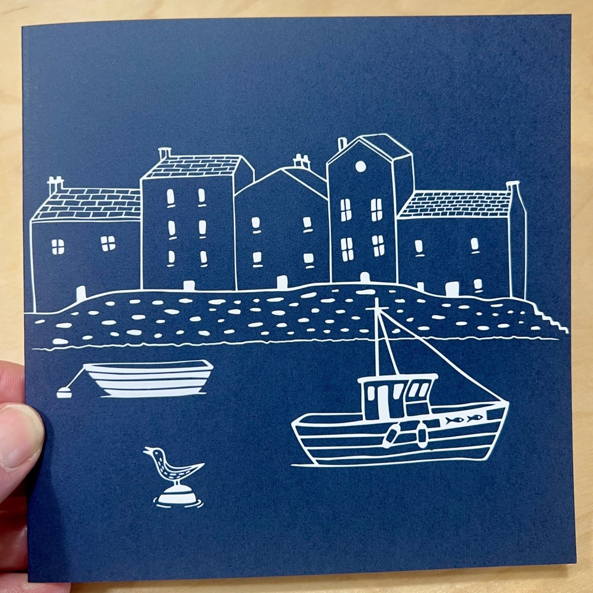 Blue card with white illustrations of houses and a boat on a wooden surface
