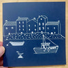 Blue card with white illustrations of houses and a boat on a wooden surface