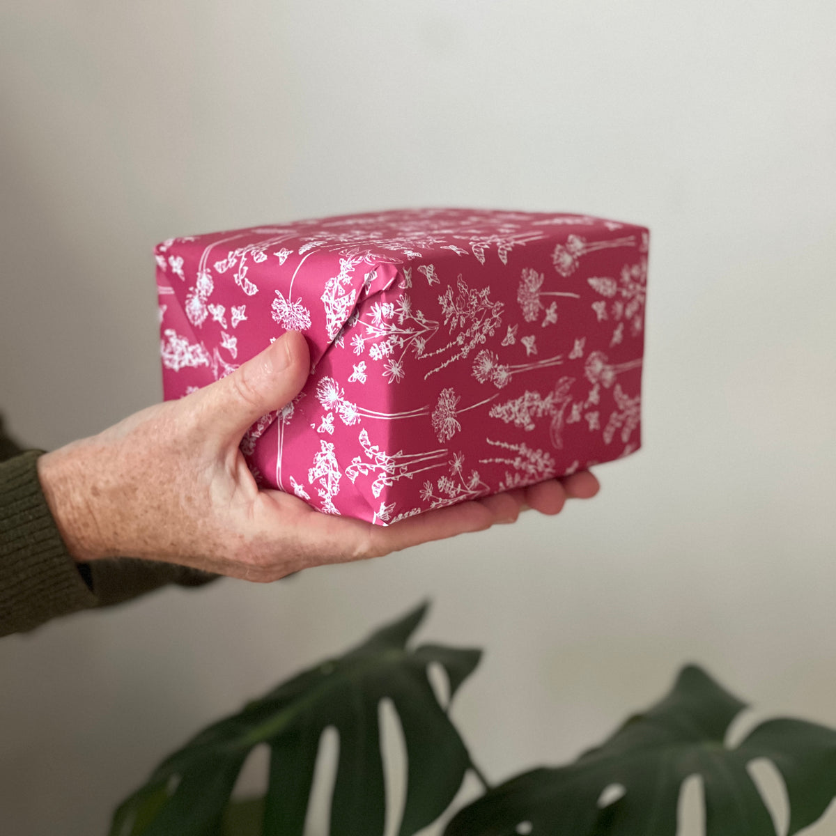 Helen is holding a square gift box wrapped in the garden raspberry red wrapping paper with a white floral pattern, set against a neutral background with a green plant leaf at the bottom.