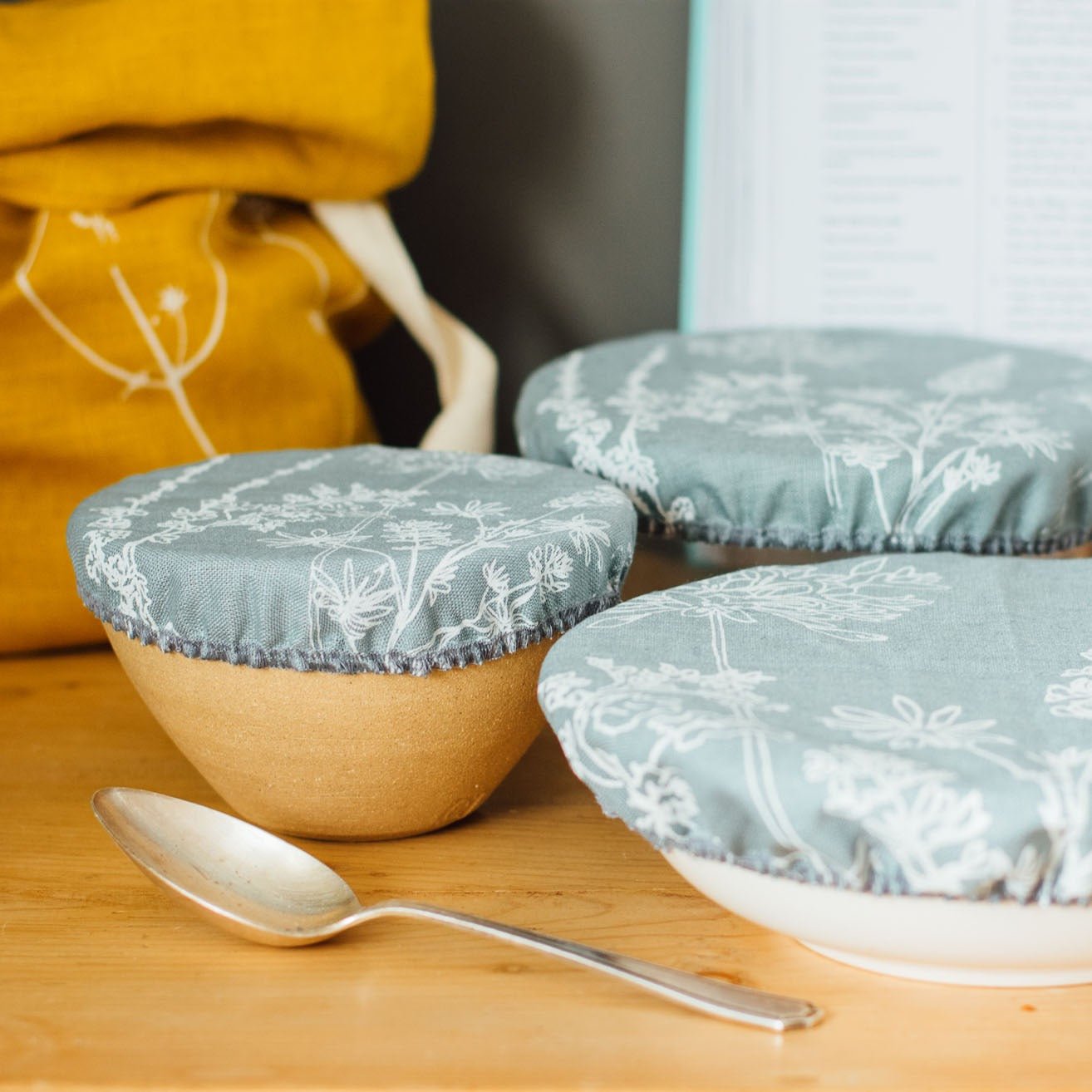 Three linen bowl covers in duck egg blue, green, and natural colours, displayed on bowls with an elasticated hem, with a spoon and a yellow bread bag in the background.