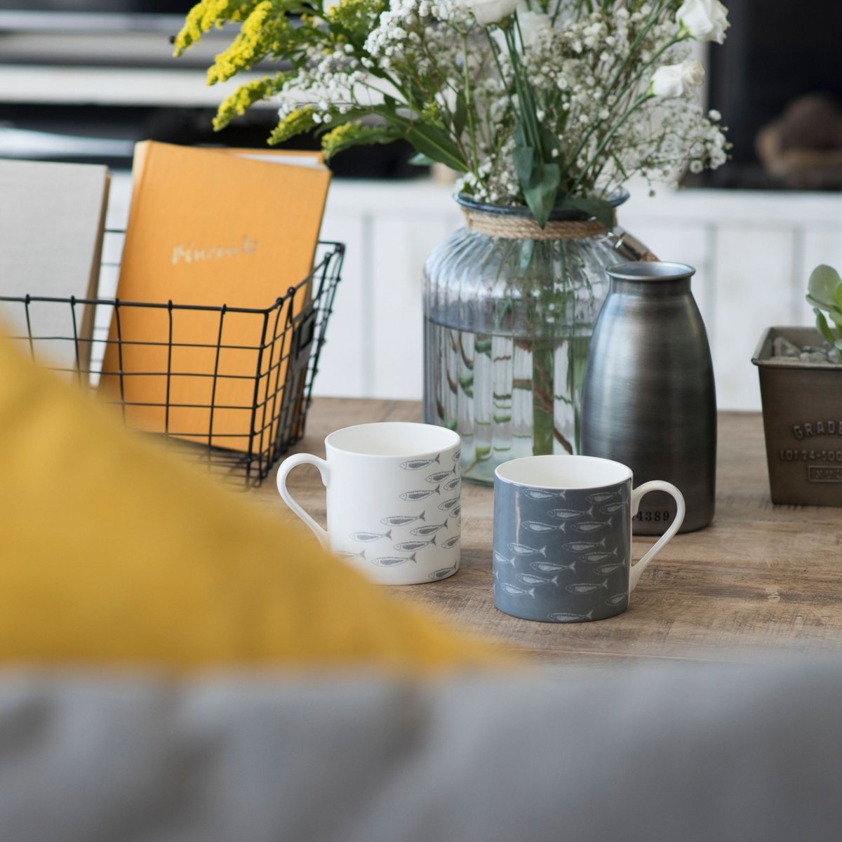 Two Fish shoal Bone China Mugs, sitting on a table, one white with grey fish, the other grey with white fish.  Both mugs have white handles  next to a vase of flowers and an empty metal vase.  Mugs from a collection by Helen Round.